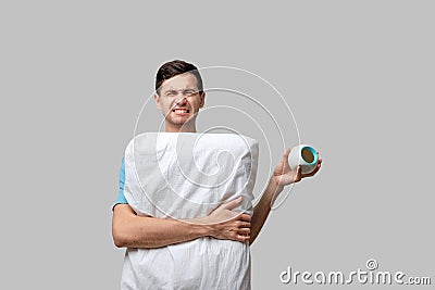 Young brunet man is upset holding alarm-clock and white pillow isolated over grey background. Stock Photo