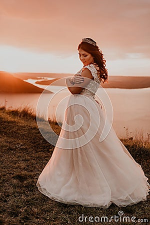 Bride in white wedding dress with a crown on her head stands on cliff against the background of the river and islands Stock Photo