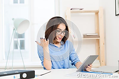 A young brunette girl in a blue shirt is sitting at the table in office. She is working with tablet. She looks plesantly Stock Photo