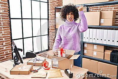 Young brunette woman with curly hair working at small business ecommerce preparing order annoyed and frustrated shouting with Stock Photo