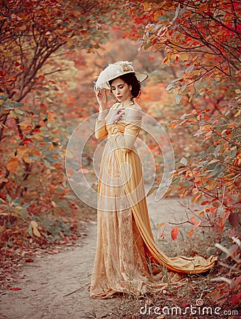 A young brunette woman with an elegant, hairstyle in a hat with a strass feathers. Lady in a yellow vintage dress walks Stock Photo