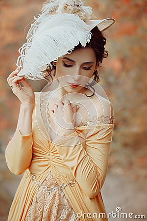 A young brunette woman with an elegant, hairstyle in a hat with a strass feathers. Lady in a yellow vintage dress walks Stock Photo