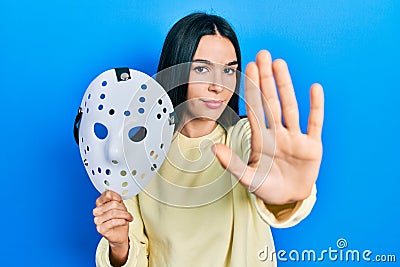 Young brunette woman holding hockey mask with open hand doing stop sign with serious and confident expression, defense gesture Stock Photo