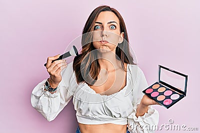 Young brunette woman holding makeup brush and blush puffing cheeks with funny face Stock Photo
