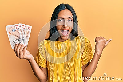 Young brunette woman holding 10 united kingdom pounds banknotes pointing thumb up to the side smiling happy with open mouth Editorial Stock Photo