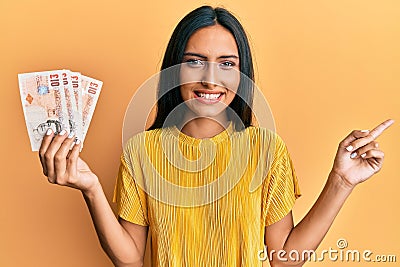 Young brunette woman holding 10 united kingdom pounds banknotes smiling happy pointing with hand and finger to the side Editorial Stock Photo