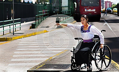 Young brunette woman sitting in wheelchair smiling with positive attitude, holding out arm looking for taxi, outdoors Stock Photo