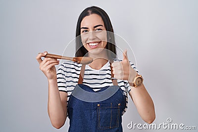 Young brunette woman wearing apron tasting food holding wooden spoon smiling happy and positive, thumb up doing excellent and Stock Photo