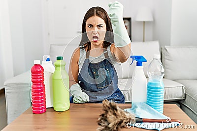 Young brunette woman wearing cleaner apron and gloves cleaning at home angry and mad raising fist frustrated and furious while Stock Photo