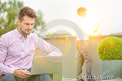 Young business man catching up on the news on his laptop was on the og outside, man reads the newspaper in the background Stock Photo