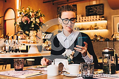 Young business woman in glasses sits in cafe at table, uses smartphone. On table is cup of coffee. Girl working Stock Photo