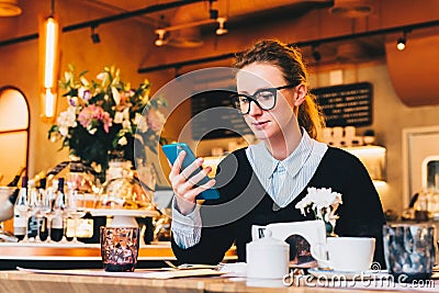 Young business woman in glasses sits in cafe at table, uses smartphone. On table is cup of coffee. Girl working Stock Photo