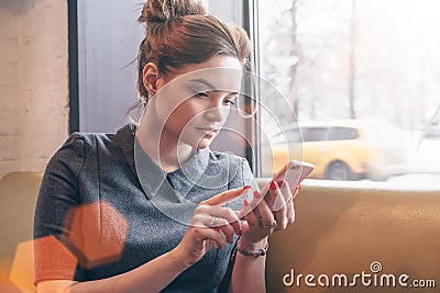 Young business woman in gray dress sitting near window and uses your smartphone.In background is blurred yellow taxi. Stock Photo