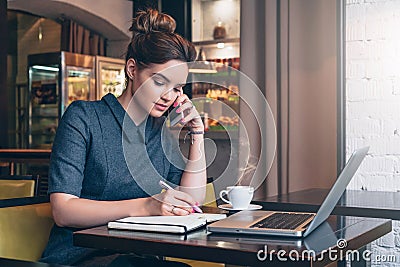 Young business woman in gray dress sitting at table in cafe, talking oncell phone while taking notes in notebook. Stock Photo