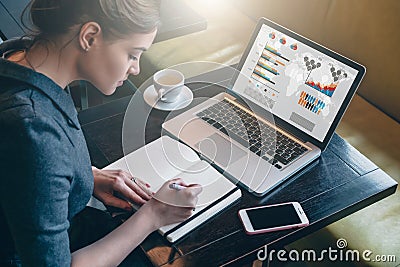 Young business woman sitting at table and taking notes in notebook.On computer screen graphics and charts. Stock Photo