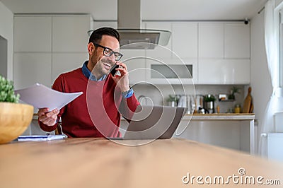 Young businessman holding tax forms and talking over mobile phone with laptop on desk in home office Stock Photo