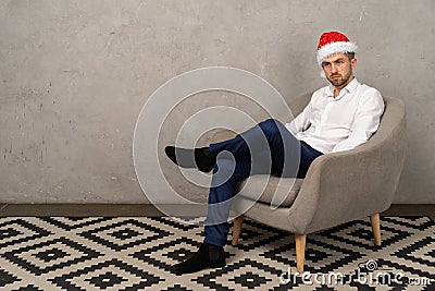Young businessman sitting on chair in santa hat while boring New Year party in the office Stock Photo