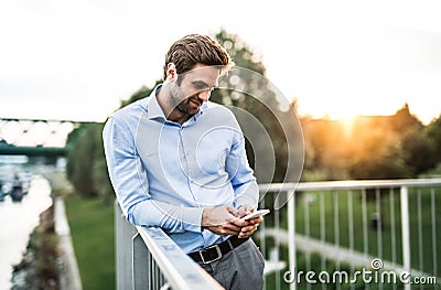 A young businessman with smartphone standing on a bridge at sunset, leaning on a railing. Stock Photo