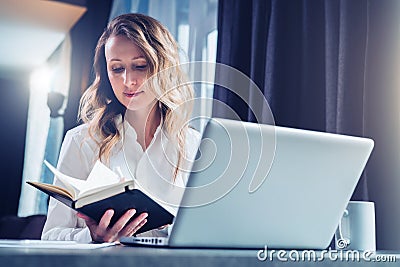 Young businesswoman in shirt is sits in office at table in front of computer and reads notes in notebook. Student Stock Photo