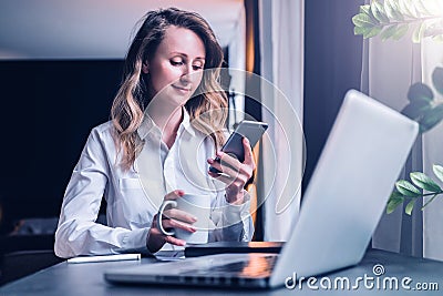 Young businesswoman in shirt is sitting in office at table in front of computer, using smartphone, looks at phone screen Stock Photo