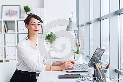 Young businesswoman sitting at her workplace, working out new business ideas, wearing formal suit and glasses, looking Stock Photo