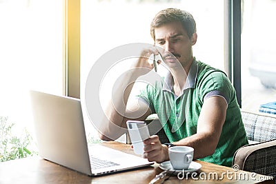 Young busy multiple businessman in green t-shirt sitting and working on laptop. talking on phone and looking at another smartphone Stock Photo