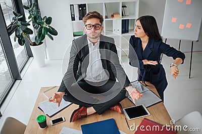 Young calm man sit at table with legs corssed. He meditate in room. Young woman get nervous. She point on watches and Stock Photo