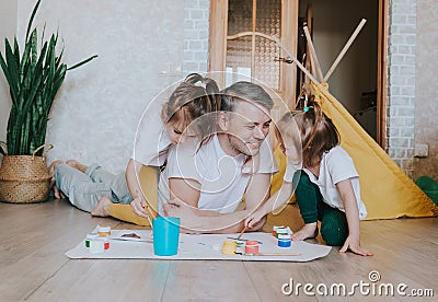 A young caring father learns to paint with his young daughters, a man lies on the floor and teaches girls to draw flowers, home Stock Photo