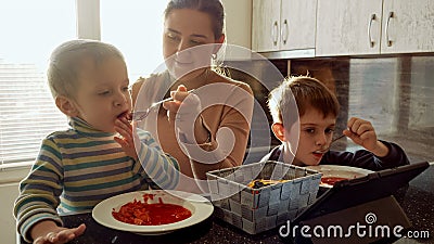 Young caring mother feeding her two sons with soup on the kitchen filled with sunset light Stock Photo