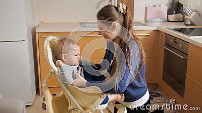 Young caring mother seating her baby son in highchair to feed him on kitchen Stock Photo