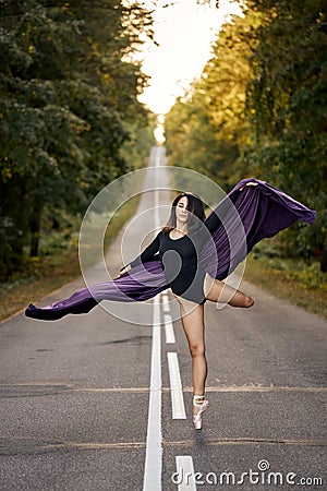 Ballerina in black bodysuit and ballet shoes dancing on road asphalt, jumps and bends Stock Photo