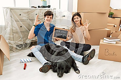 Young caucasian couple with dog holding our first home blackboard at new house smiling positive doing ok sign with hand and Stock Photo