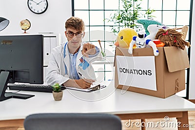 Young caucasian doctor man working at the clinic with toys donations box for children pointing with finger to the camera and to Stock Photo