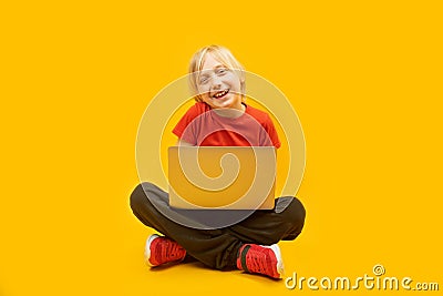 Young caucasian fair-haired student sits on the floor and holds laptop. Boy study at home with internet and computer on yellow Stock Photo