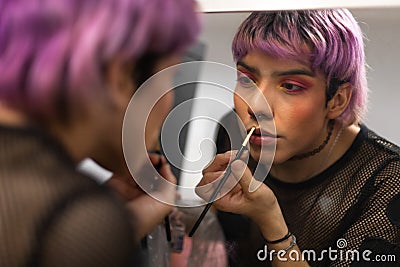 Young caucasian male with purple hair applying lipstick with make up brush looking in the mirror. Stock Photo