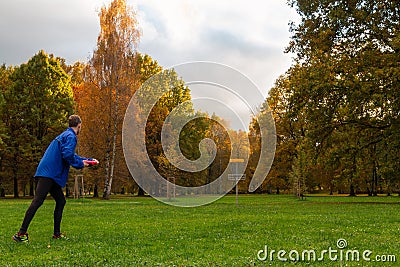 Young caucasian man in blue jacket playing disc golf on autumn play course with basket Stock Photo