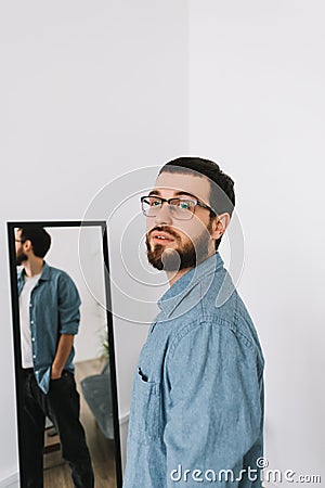 Young caucasian man in glasses standing at the mirror in living room Stock Photo