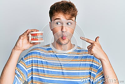 Young caucasian man holding denture making fish face with mouth and squinting eyes, crazy and comical Stock Photo