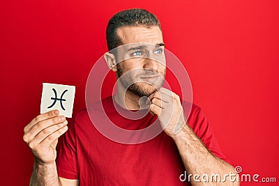 Young caucasian man holding paper with pisces zodiac sign serious face thinking about question with hand on chin, thoughtful about Stock Photo