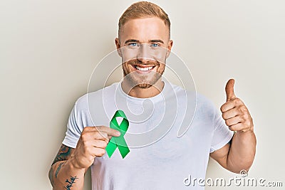 Young caucasian man holding support green ribbon smiling happy and positive, thumb up doing excellent and approval sign Stock Photo