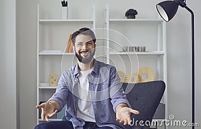 Young caucasian man looking at camera video chat with open arms in friendly welcome gesture in living room. Stock Photo