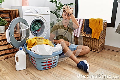 Young caucasian man putting dirty laundry into washing machine surprised with hand on head for mistake, remember error Stock Photo