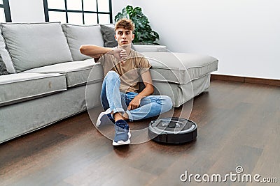 Young caucasian man sitting at home by vacuum robot with angry face, negative sign showing dislike with thumbs down, rejection Stock Photo