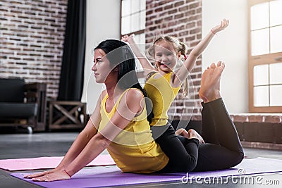 Young Caucasian mother doing yoga cobra pose on floor while her smiling daughter sitting on moms back Stock Photo