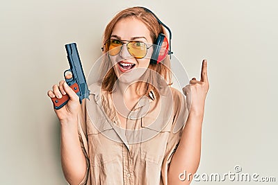 Young caucasian shooter woman using gun smiling with an idea or question pointing finger with happy face, number one Stock Photo