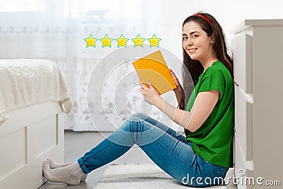 A young Caucasian woman sits on the floor and shows a book with an excellent rating, a hit of sales. Bright room in the background Stock Photo