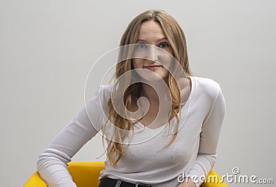 Young caucasian woman interested in coversation and looking at camera while sitting in chair, grey background Stock Photo