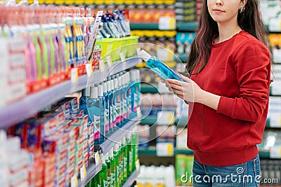 A young Caucasian woman in a red sweater holds a bottle of shampoo and smiles tenderly. In the background, shelves with cosmetics Stock Photo