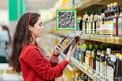 A young Caucasian woman in a red sweatshirt scans the qr code on a bottle of oil using smartphone. Qr code icon above the product Stock Photo