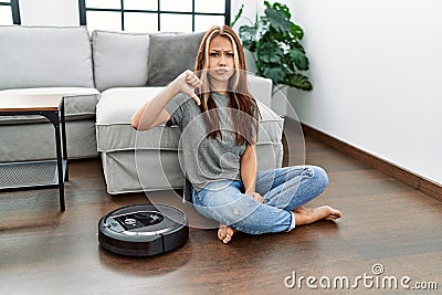 Young caucasian woman sitting at home by vacuum robot with angry face, negative sign showing dislike with thumbs down, rejection Stock Photo